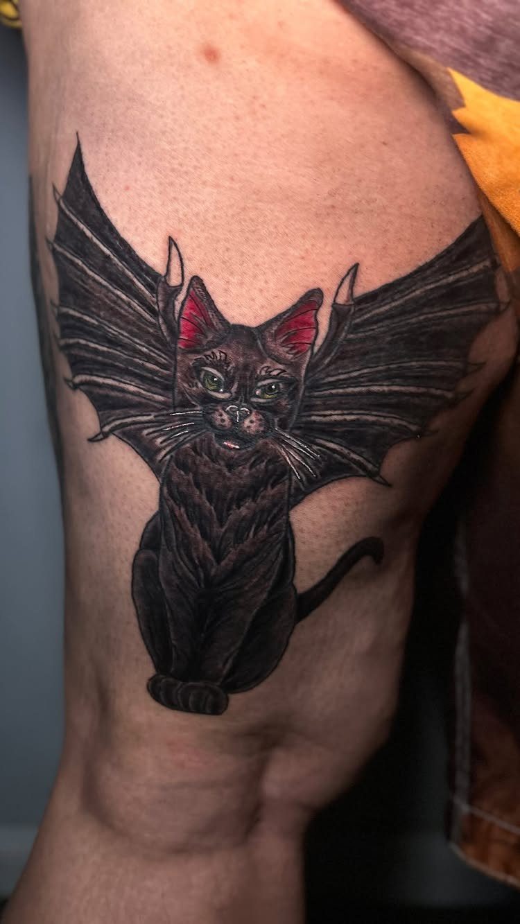 Black cat tattoo with bat wings and red ears on a person's leg