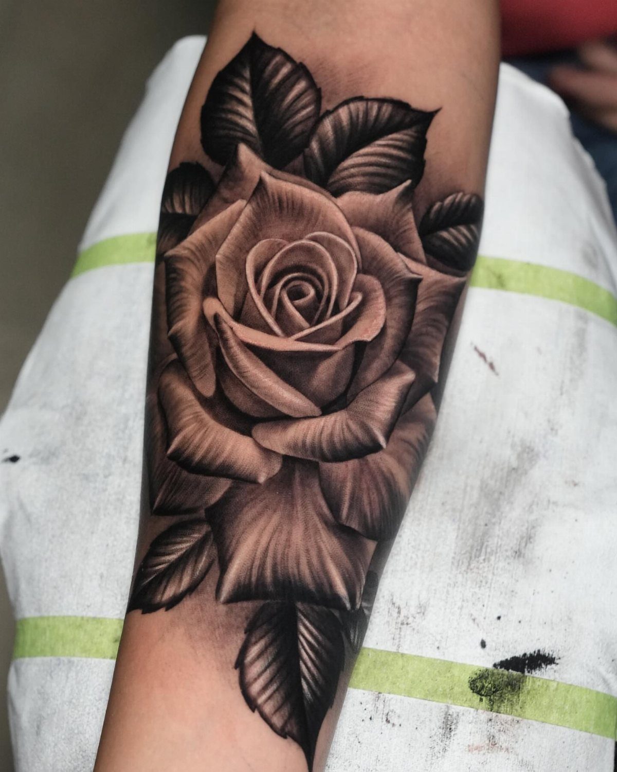 Realistic rose bloom tattoo with detailed shading and dark leaves on a forearm.
