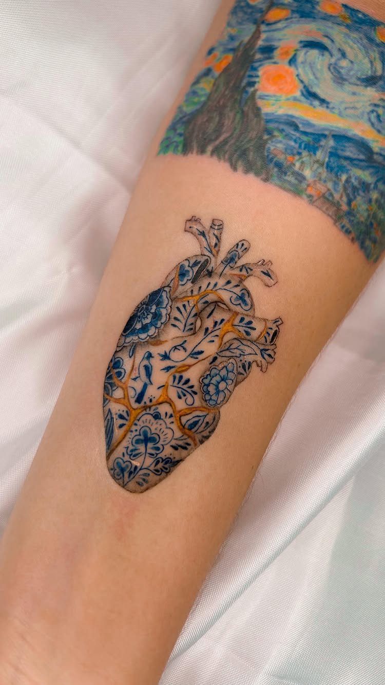 Anatomical heart tattoo with blue floral patterns and gold kintsugi cracks on skin.
