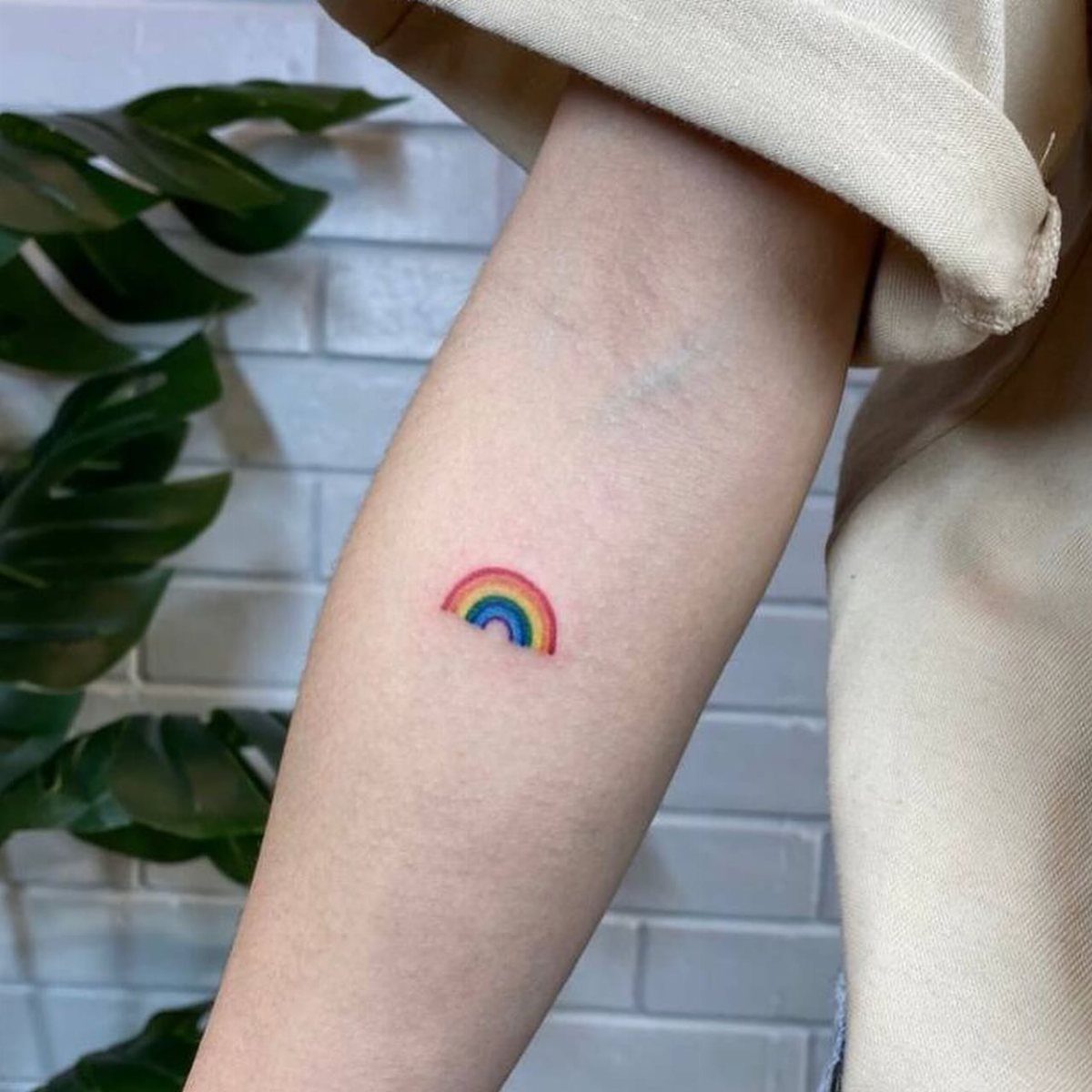 Small colorful rainbow tattoo on a person's inner forearm against a white brick wall.