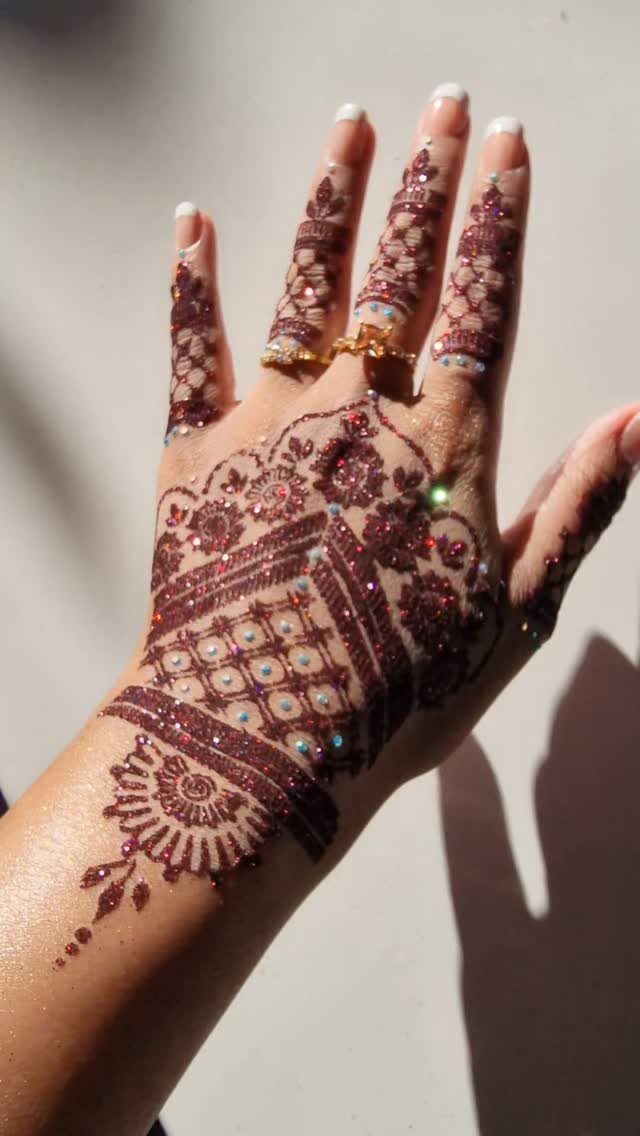 Hand with intricate reddish-brown henna tattoo decorated with sparkling multicolored glitter accents.