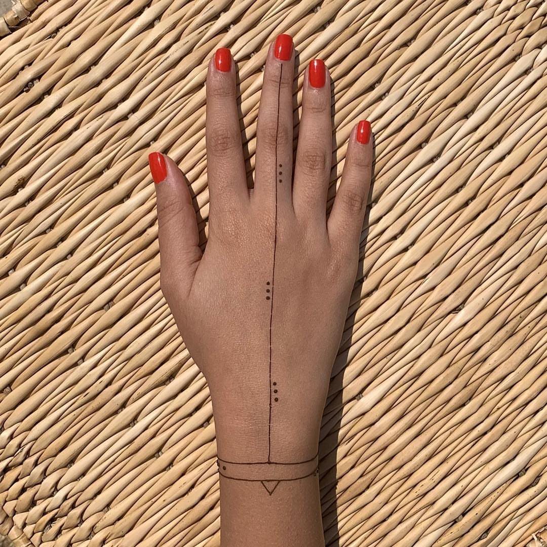 Hand with red nails and a minimal henna line and dot tattoo on straw.