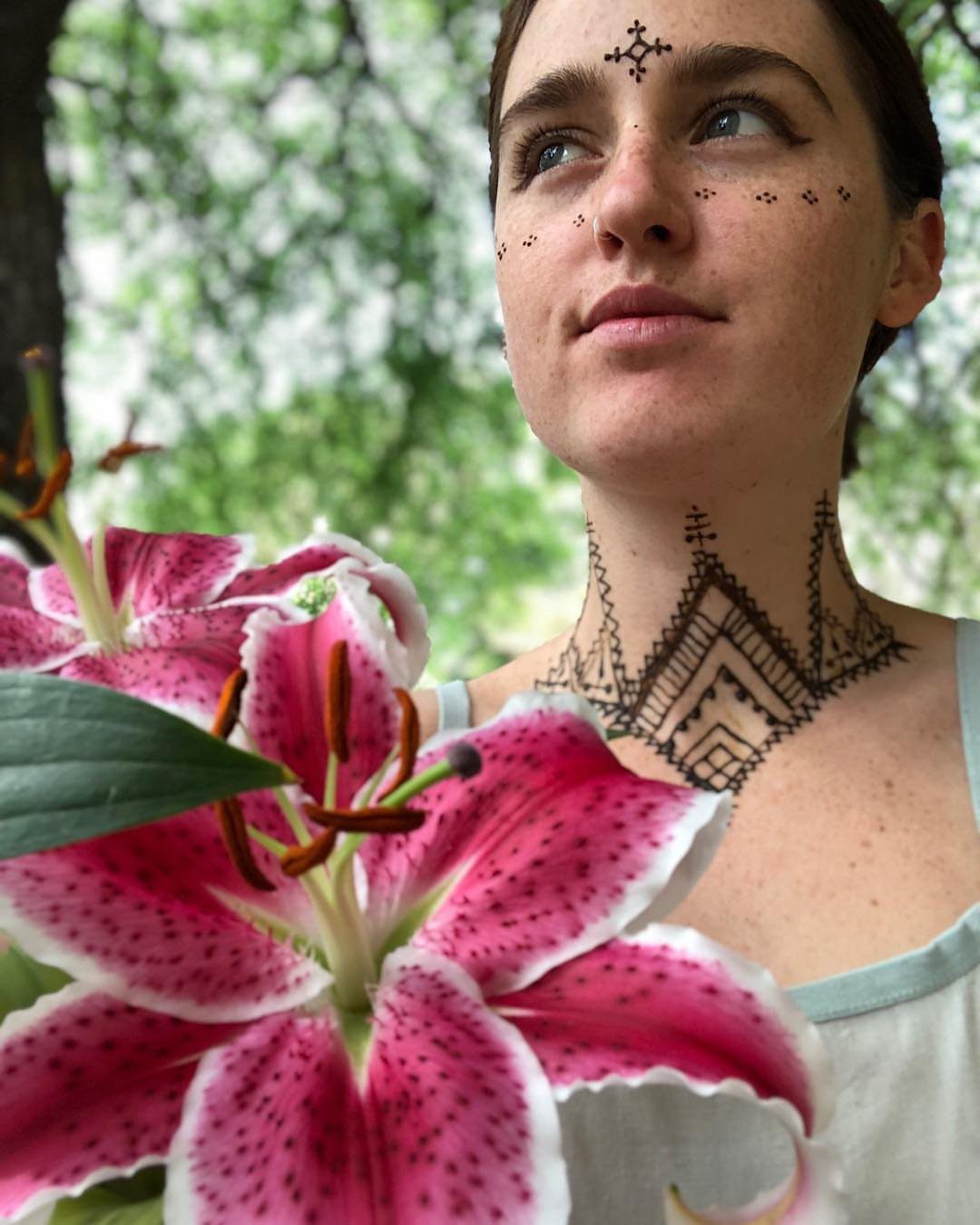Woman with intricate dark henna tattoos on her neck and face near pink lilies.