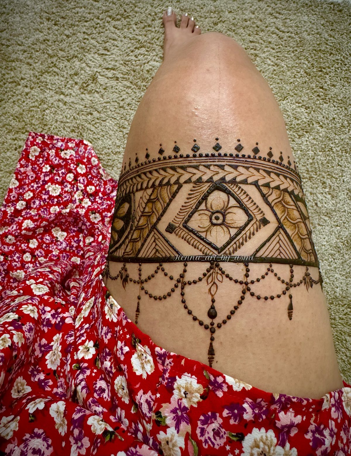Intricate brown henna tattoo design on a person's thigh with red floral clothing.