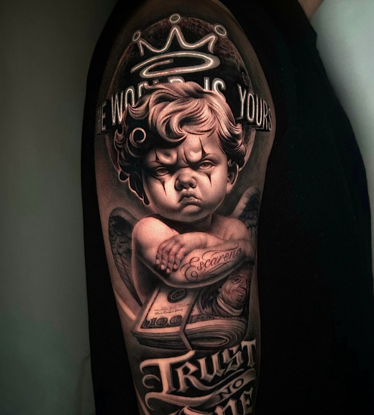 Black and grey realism tattoo of a frowning cherub with money and text.