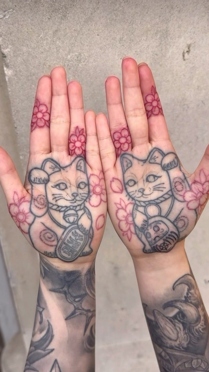 Two palms with Japanese lucky cat tattoos and red cherry blossoms on fingers.
