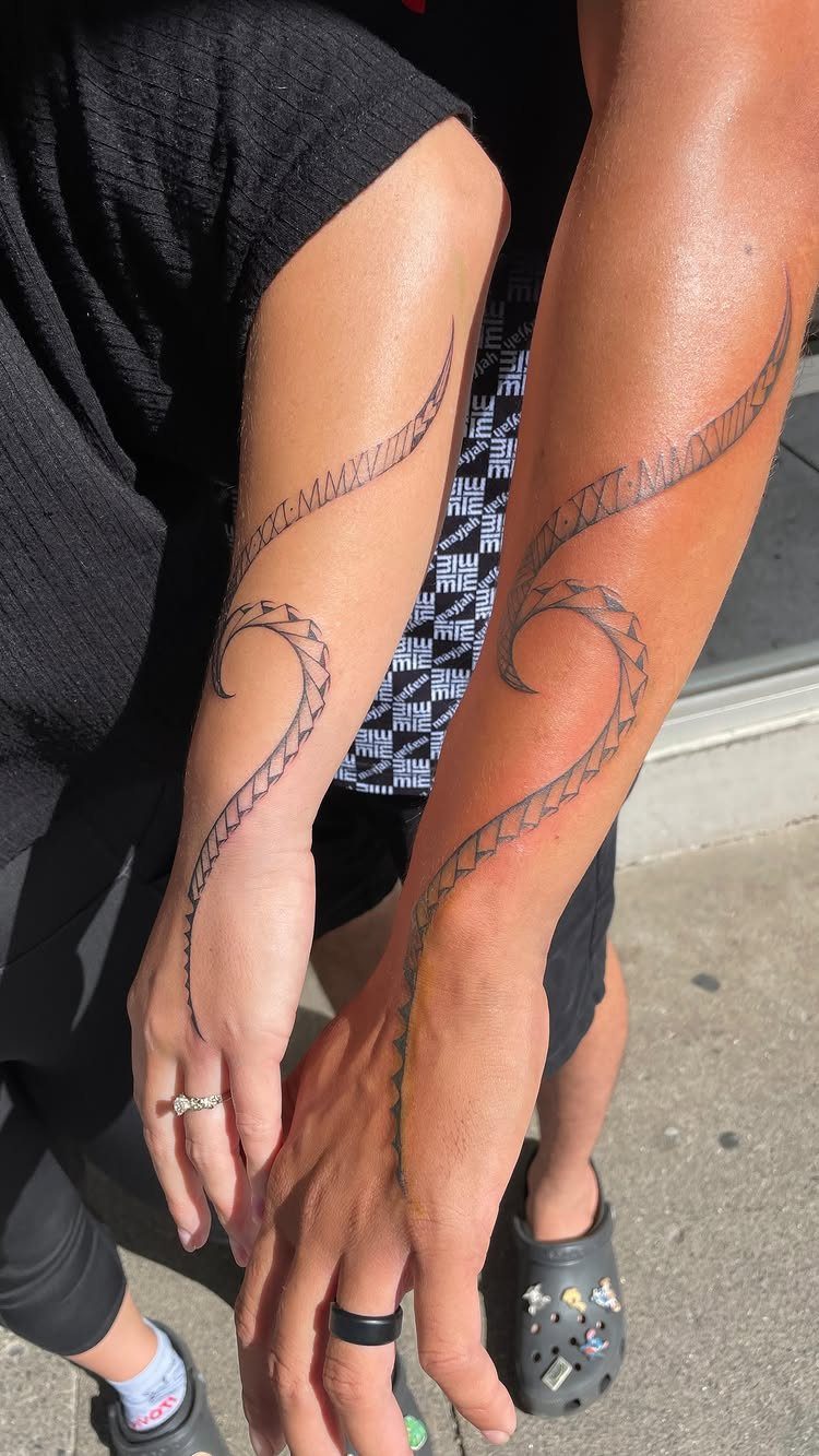 Matching black ink swirling tribal tattoos on two people's forearms outdoors.