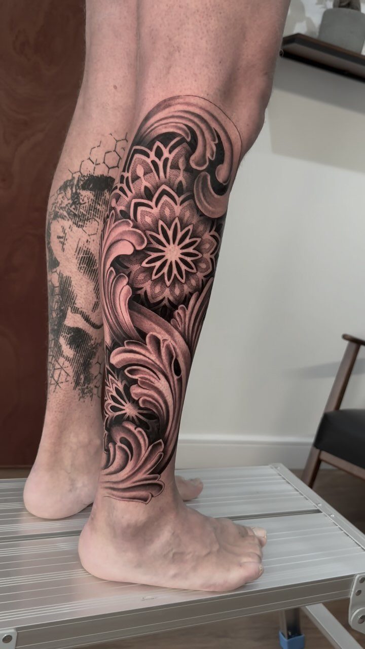 Detailed black and grey mandala tattoo with ornate scrolls on a lower leg.