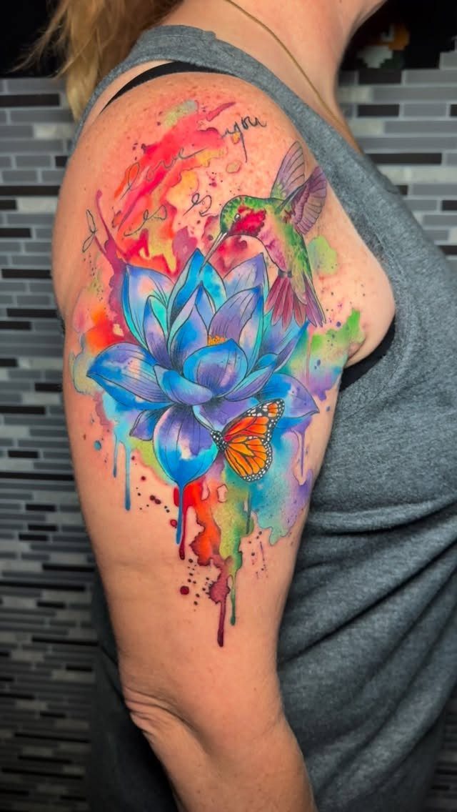 Watercolor tattoo on arm featuring a blue lotus, hummingbird, and orange butterfly.