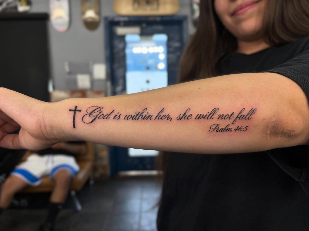 Forearm tattoo featuring a small cross and a biblical verse from Psalm 46:5