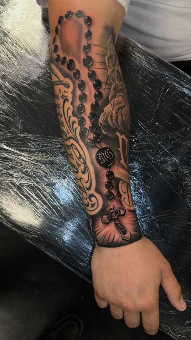 Rosary with cross tattoo on a forearm with clouds and script lettering.