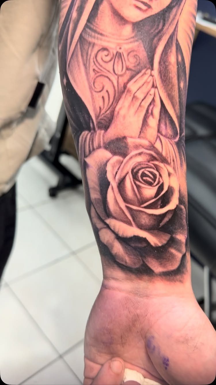 Virgin Mary tattoo with praying hands and a rose on a forearm.