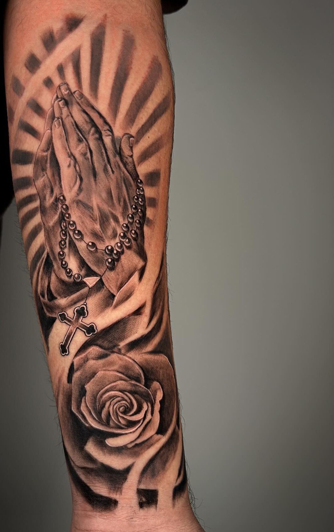 Praying hands with rosary beads and a rose flower tattoo on an arm.