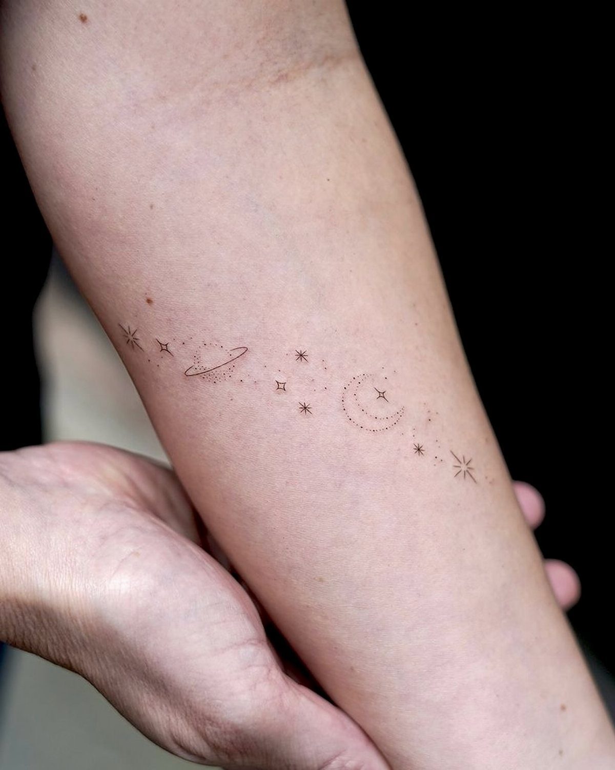 Thin line tattoo of a moon, stars, and planet on a forearm