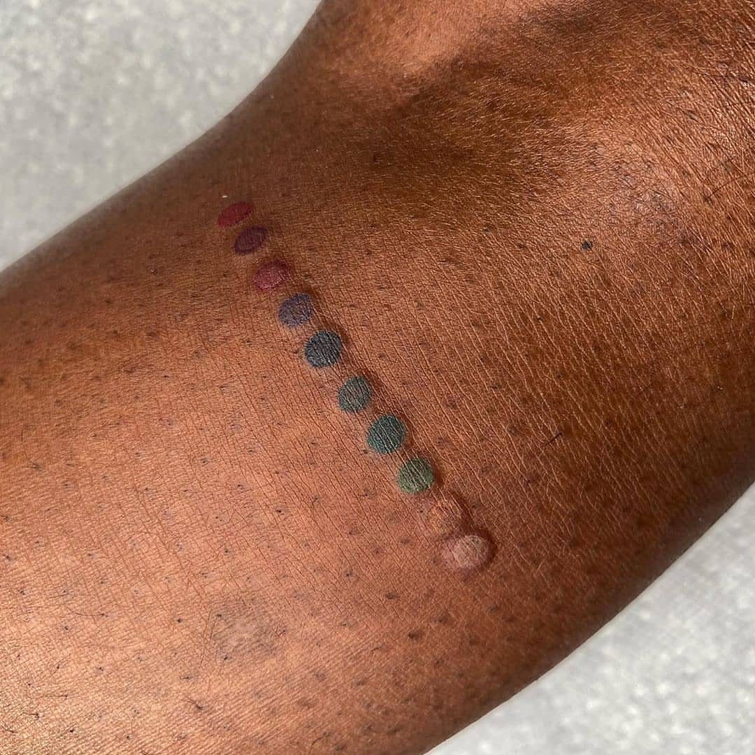 Row of colorful ink dots on dark brown skin for tattoo color testing.