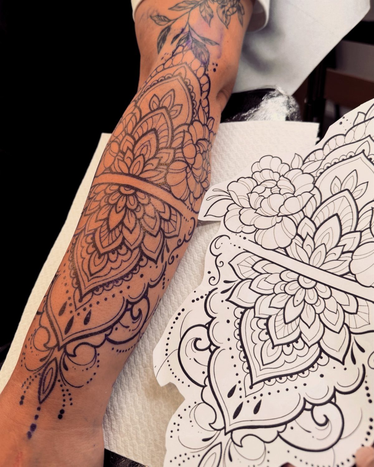 Floral and mandala tattoo stencil applied to a forearm next to paper design.