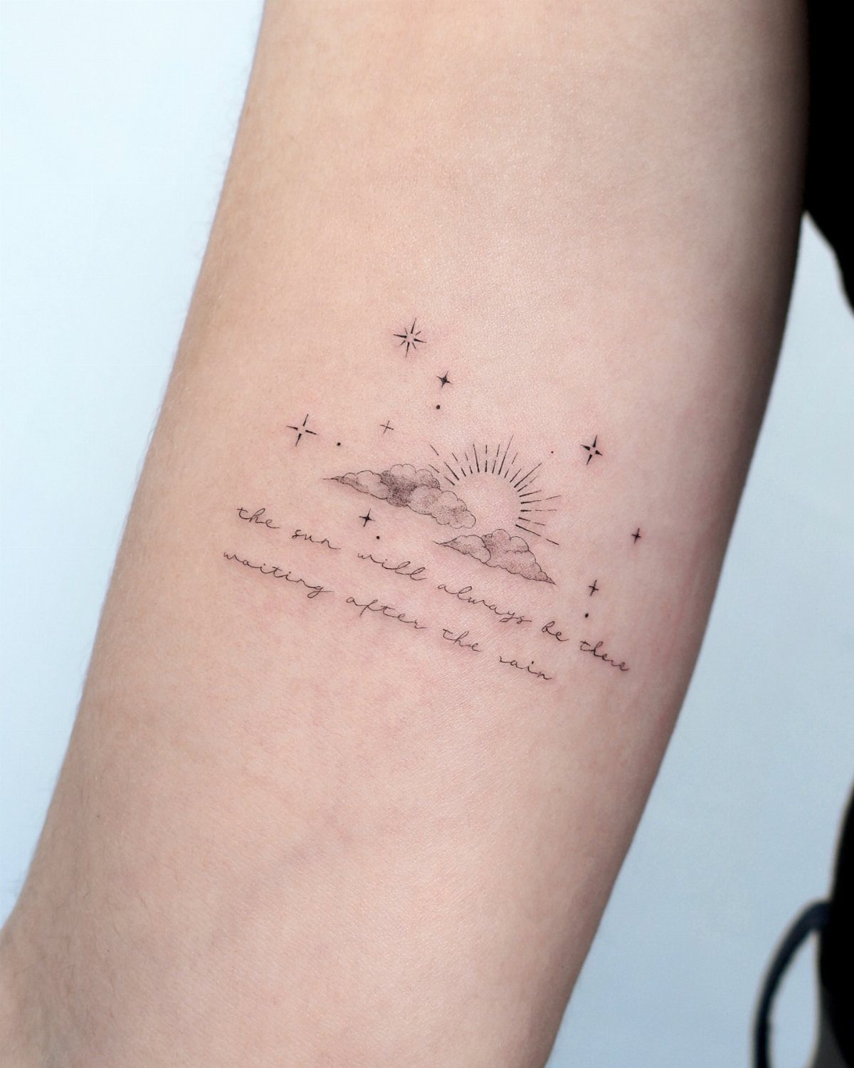 Tattoo on a forearm showing small stars, clouds, a sun, and cursive text.