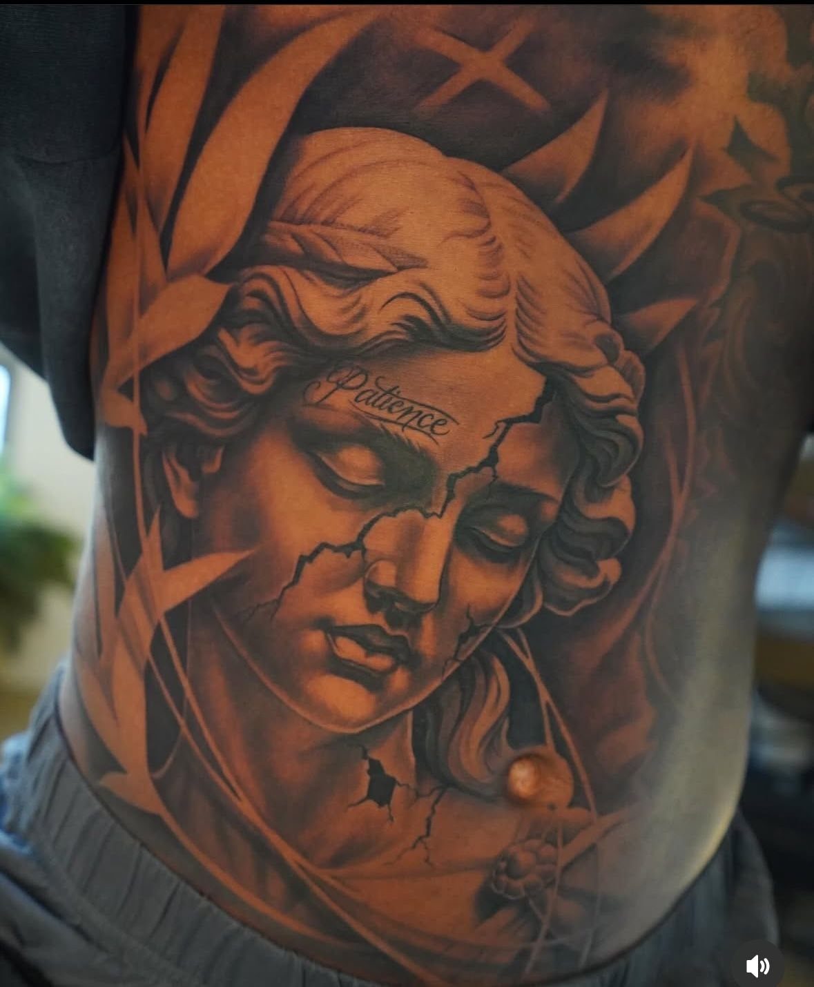 Realistic black and grey tattoo of a cracked statue face on a stomach.