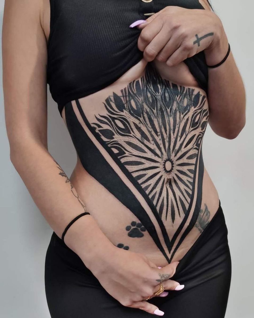 Geometric black ink tattoo with symmetrical radiating patterns covering a person's stomach area.