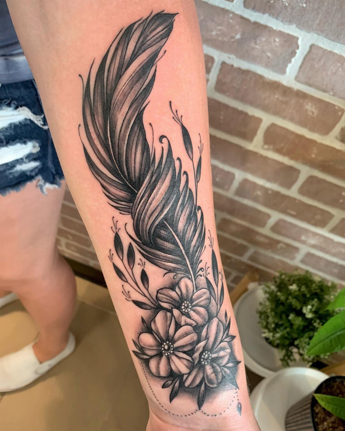 Feather and flower tattoo in black and grey ink on a person's forearm.