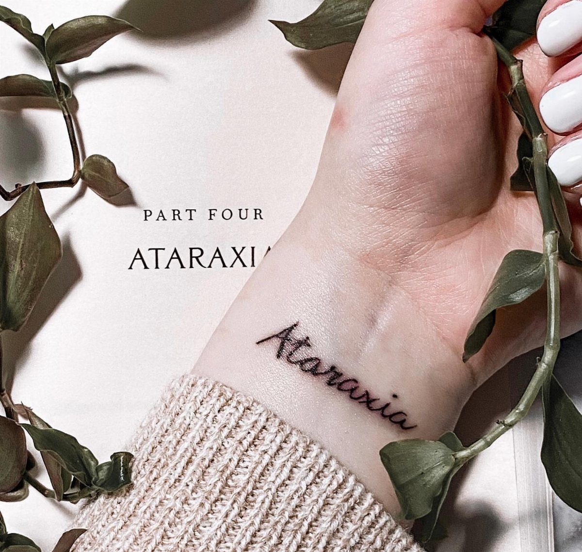 Cursive Ataraxia tattoo on a wrist resting on a book with green leaves.