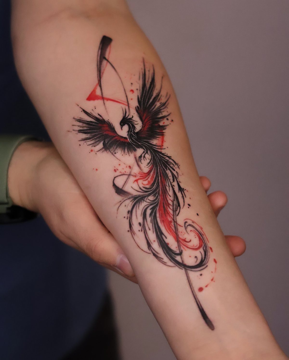 Phoenix tattoo on a forearm featuring black and red ink with flowing feathers.