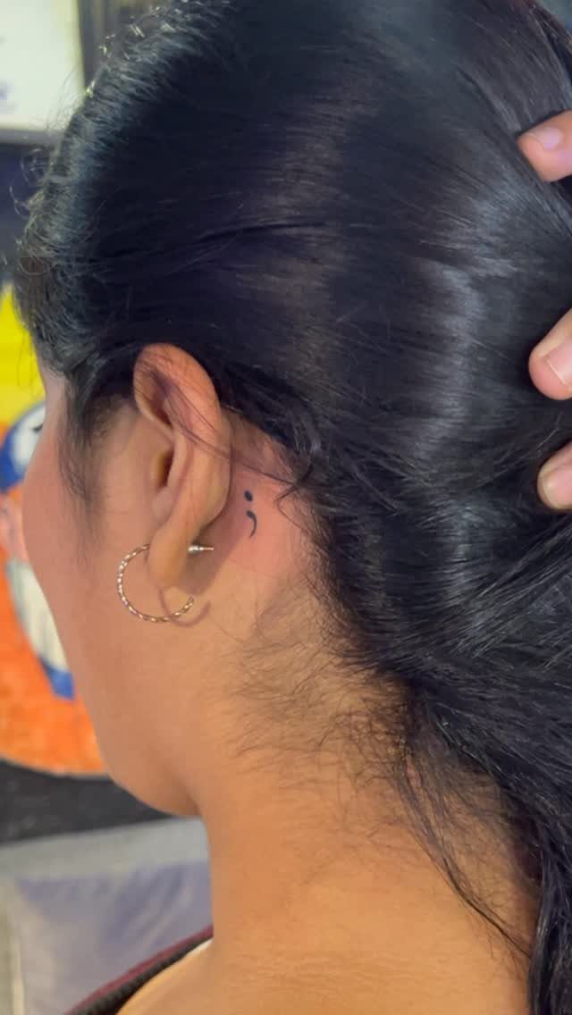 Small black semicolon tattoo located behind the ear of a person with black hair.