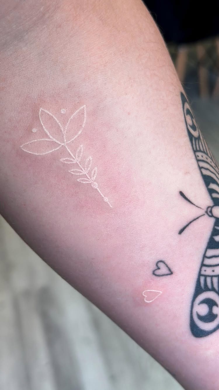 White ink flower and heart tattoos on a person's arm next to a dragonfly.