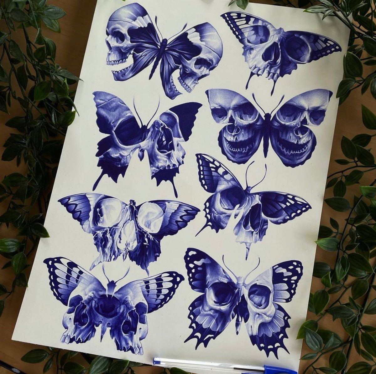 Blue ink tattoo stencils of skulls combined with butterfly wings on white paper.