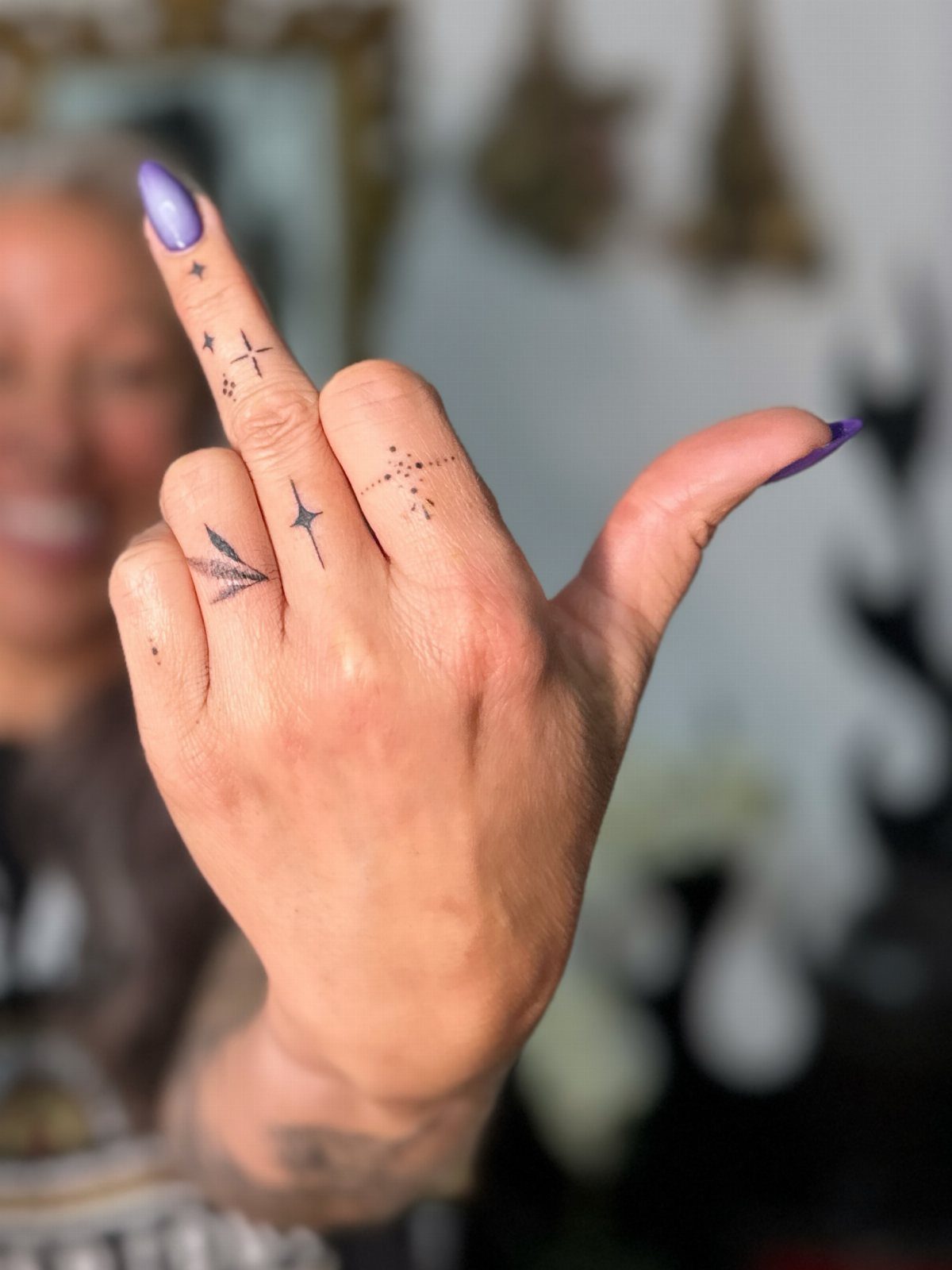 Hand with purple nails featuring small brown geometric tattoos resembling henna designs.