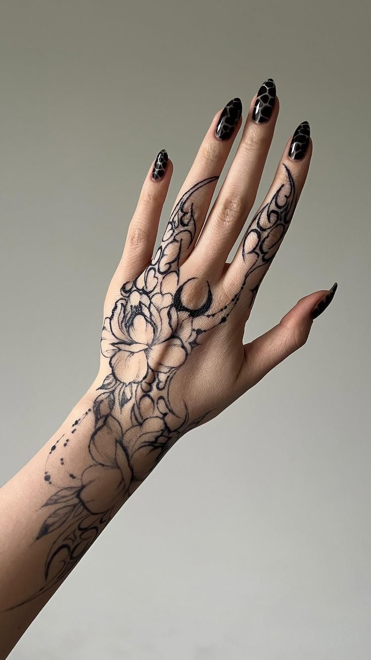 Hand with a black floral and moon tattoo and patterned black nails.
