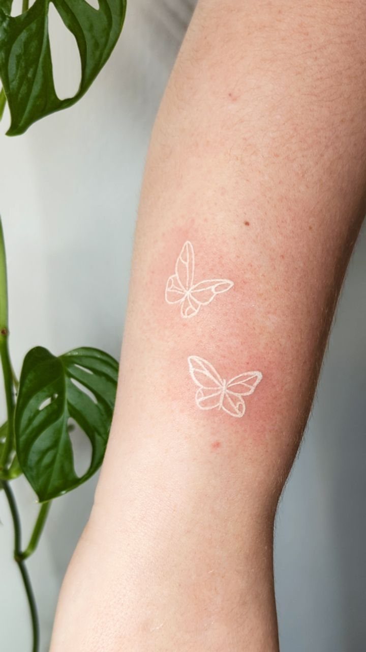 Two white ink butterfly tattoos on a person's forearm next to green leaves.