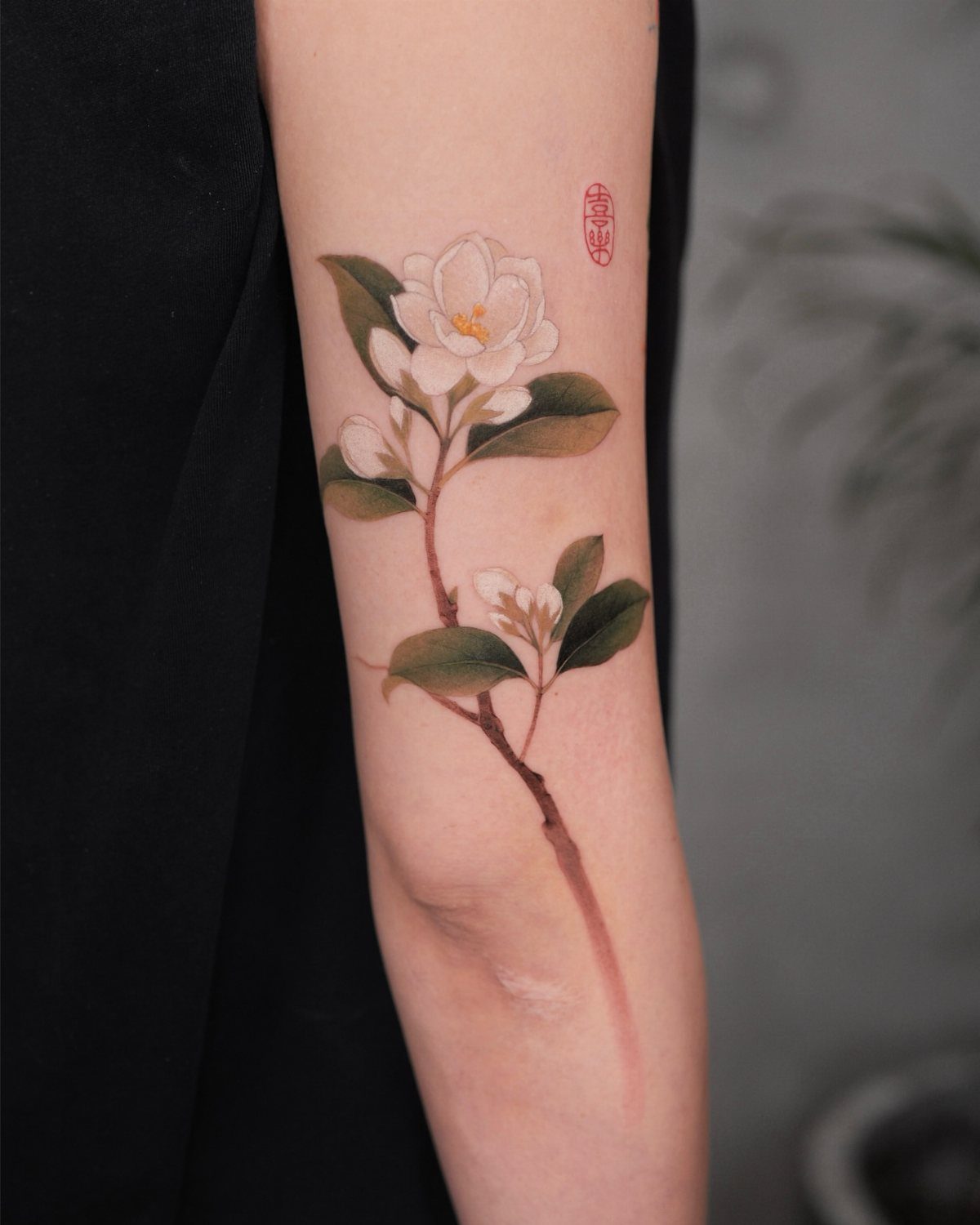 White flower tattoo with green leaves and a brown stem on a forearm.