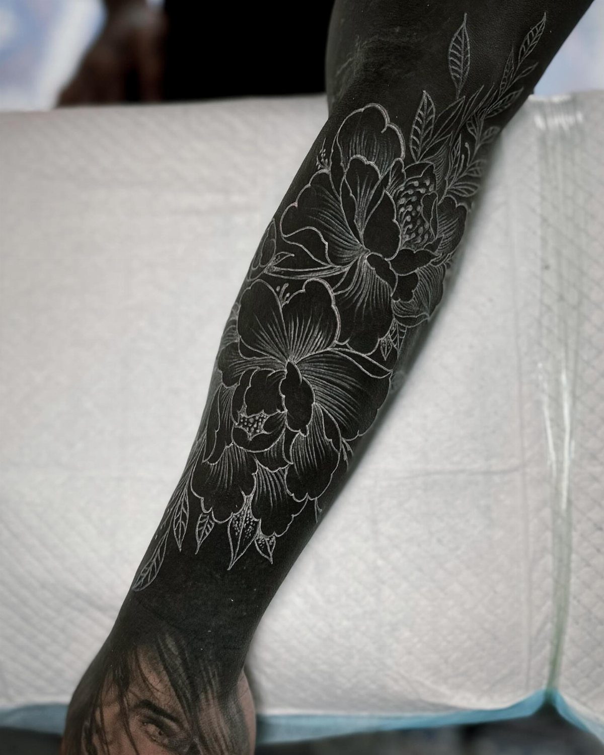 White ink peony flowers tattooed over a solid black ink sleeve on an arm.