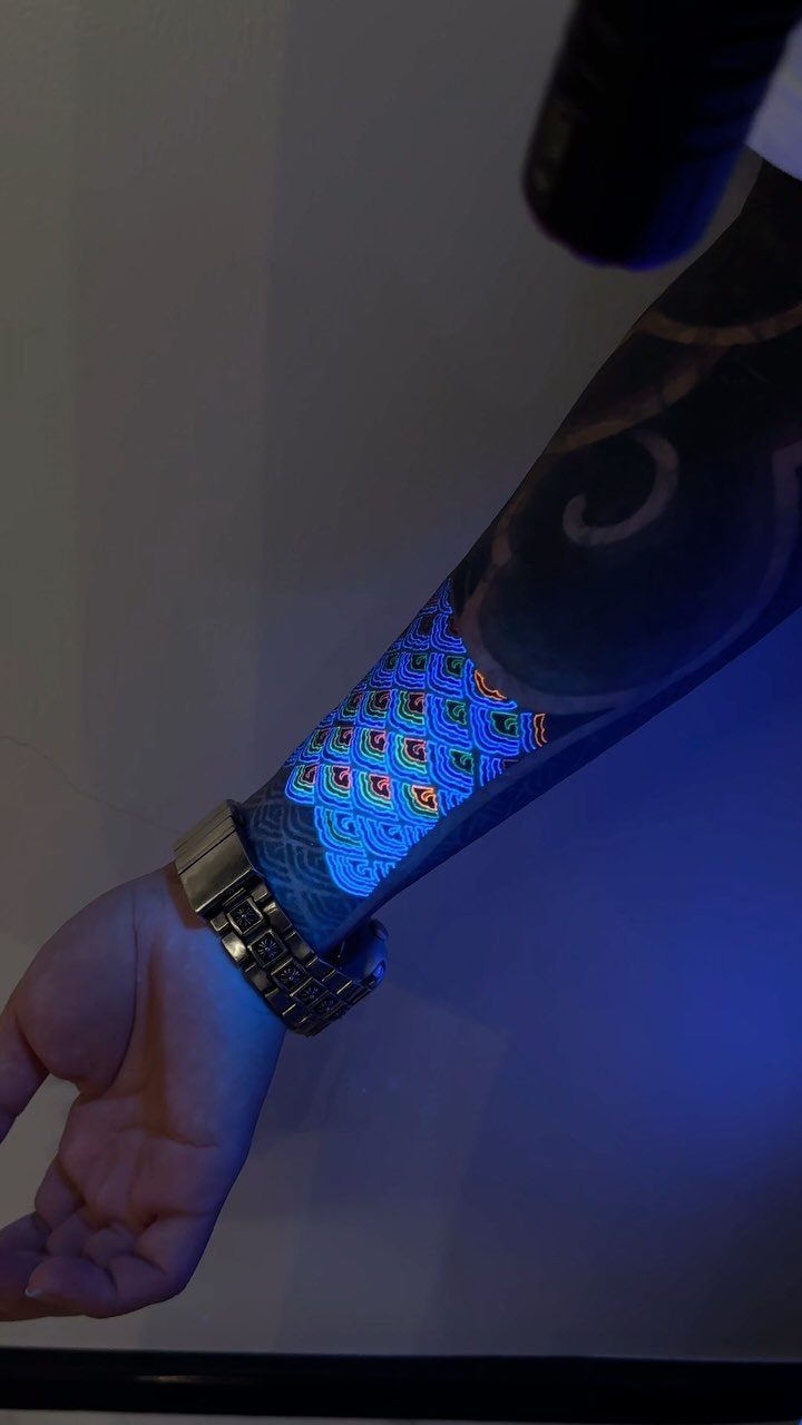 Glowing neon scale tattoo on a forearm under a blacklight with a silver watch.