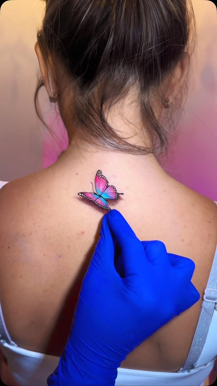 Colorful butterfly tattoo on a person's upper back, pointed to by a blue glove.