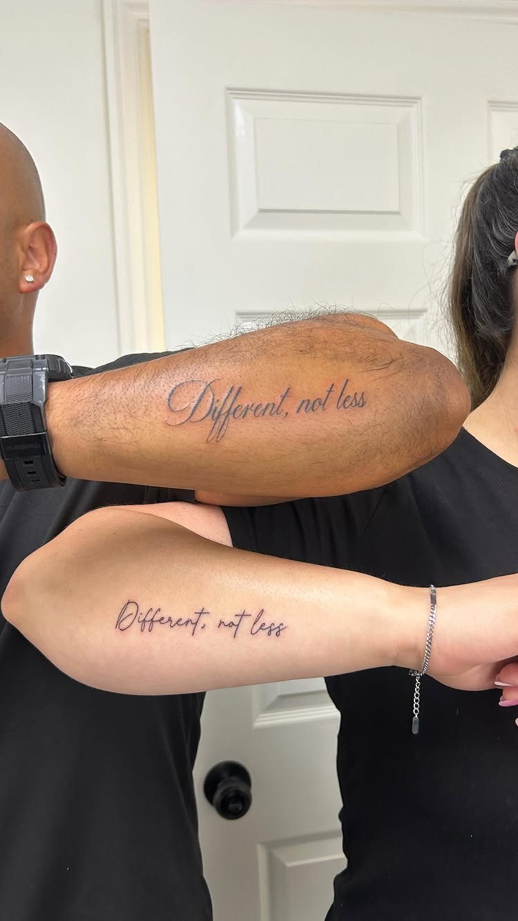 Two forearms with matching cursive tattoos reading Different, not less against a white door.
