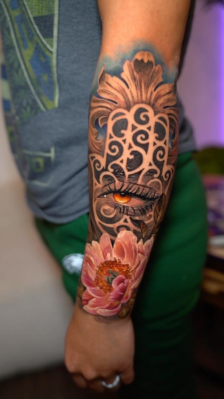 Hamsa hand tattoo with an eye and pink lotus flower on a forearm.