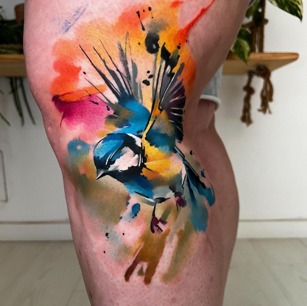 Watercolor tattoo of a blue bird with colorful ink splashes on a thigh.