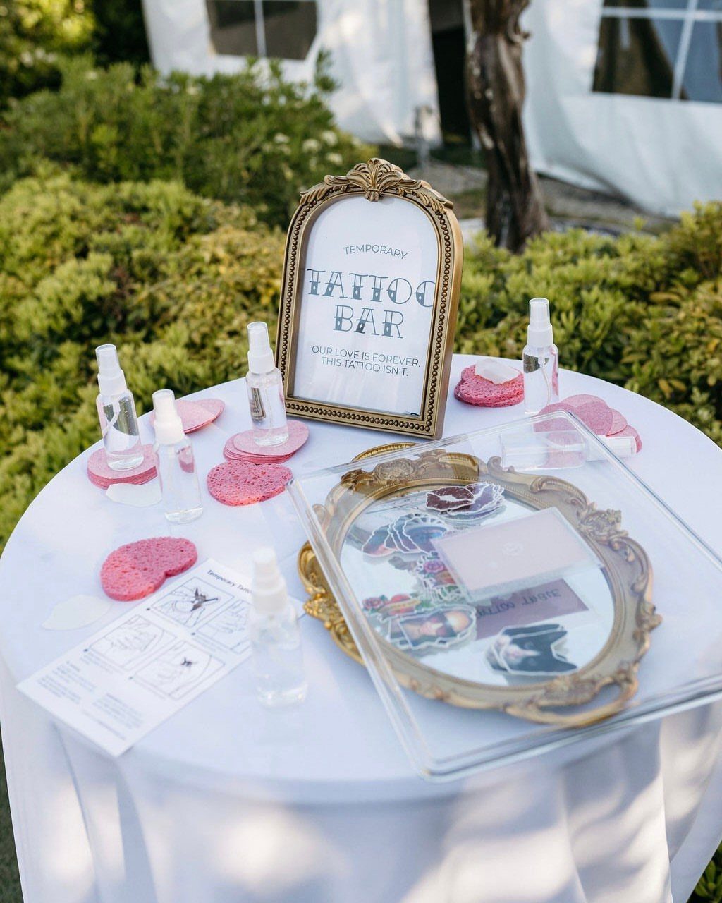 Temporary tattoo bar setup with gold tray, spray bottles, and pink sponges on white table.