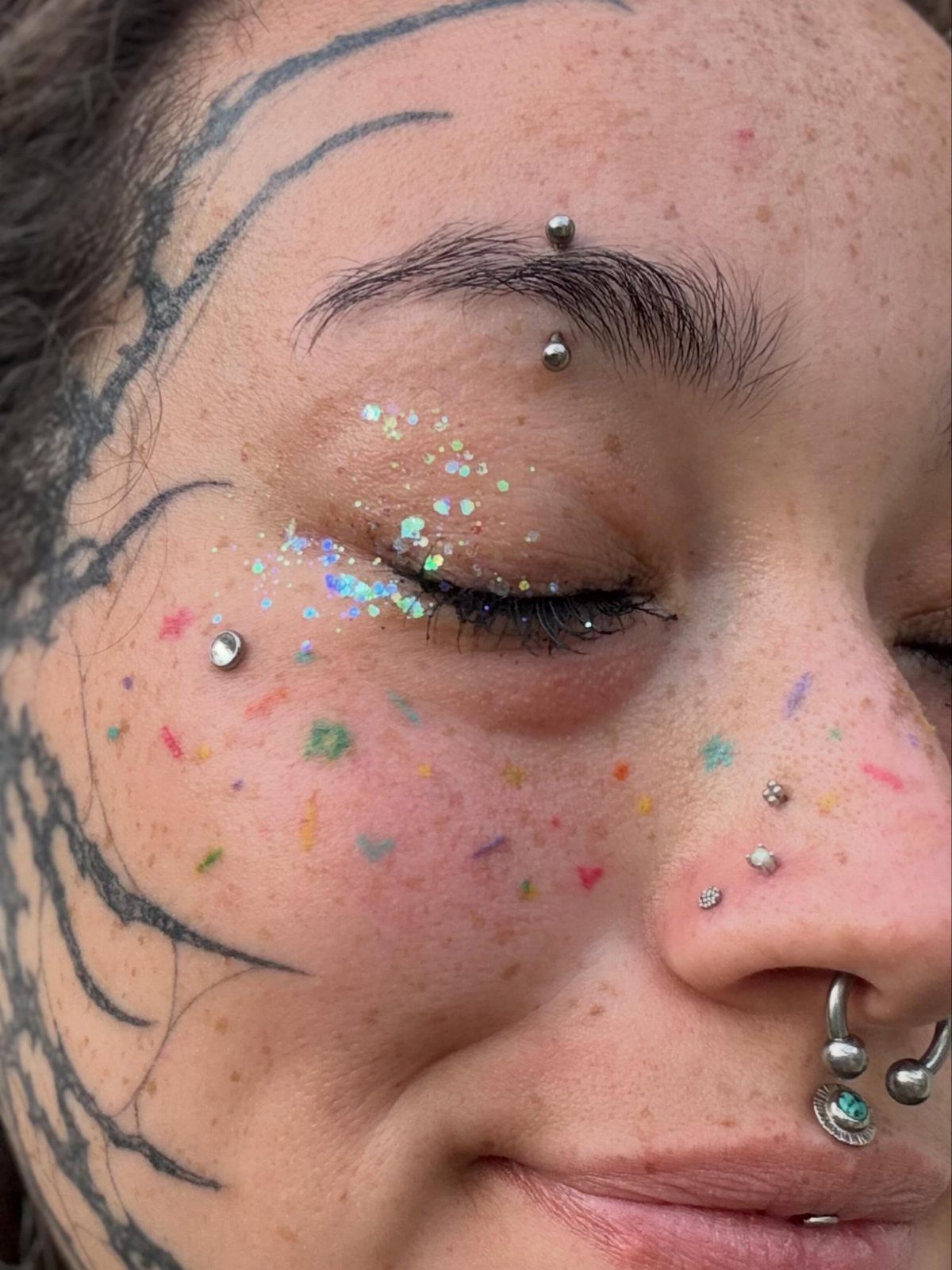 Person's face with black ink tattoos, colorful glitter makeup, and various facial piercings.
