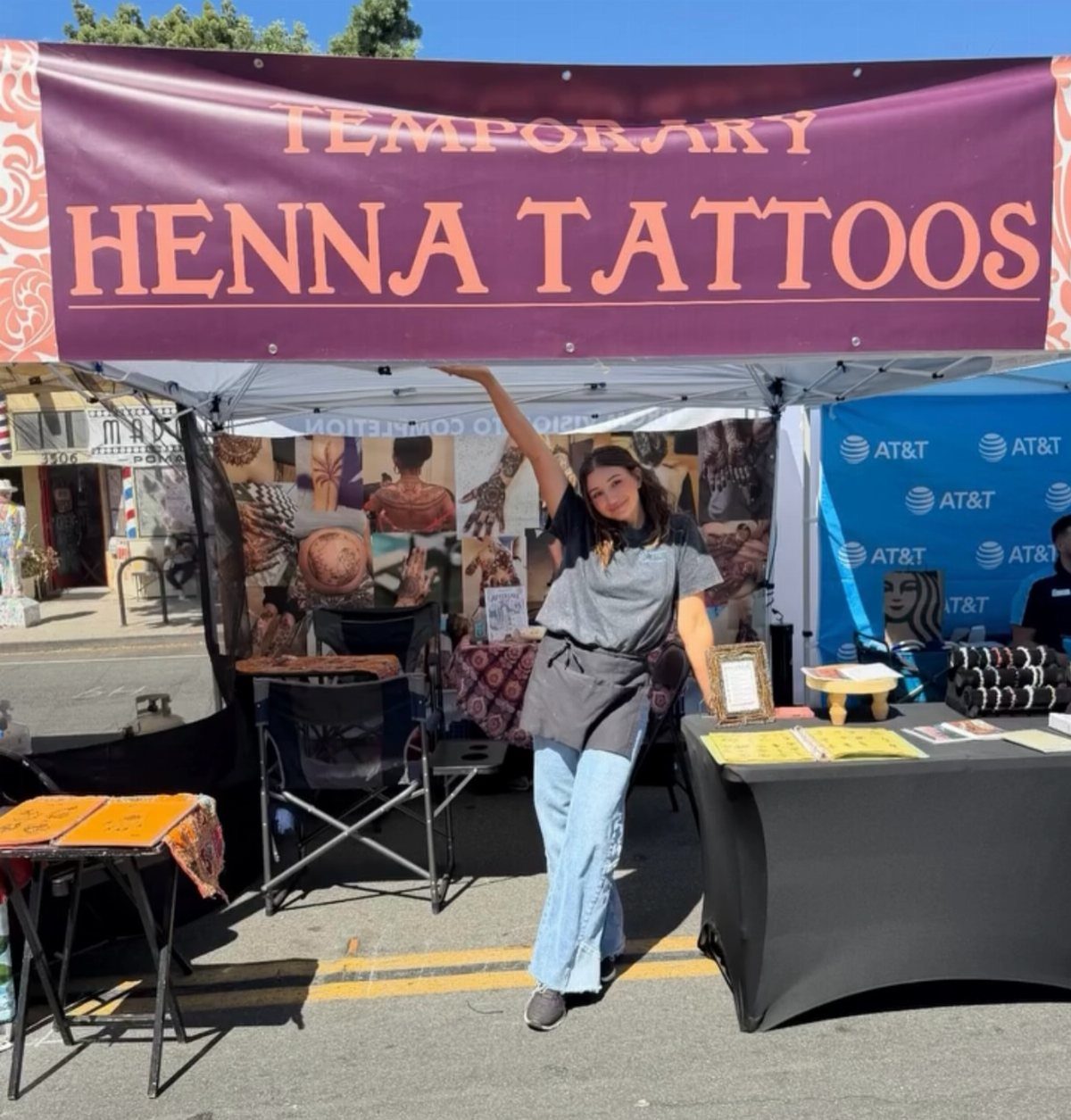 Henna tattoo booth setup with a purple banner and a woman standing by a table.