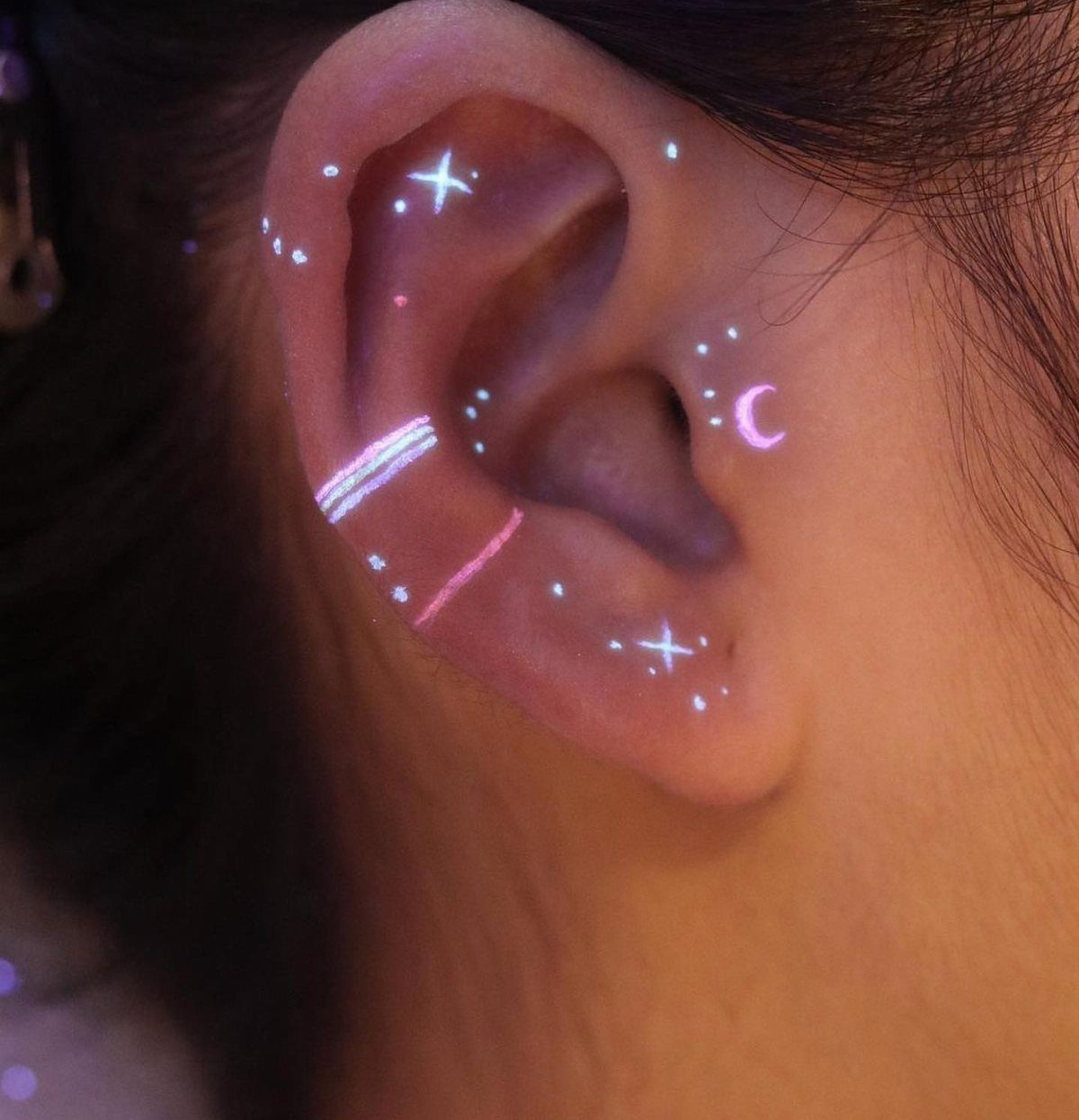 Glowing purple and white UV tattoos of stars and a moon on an ear.