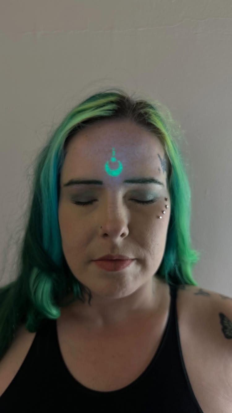Person with green hair showing a glowing blue UV crescent moon forehead tattoo.
