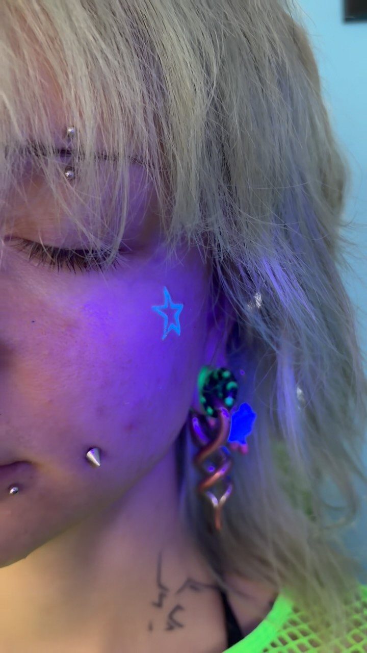 Person with blonde hair and a glowing blue star UV face tattoo.