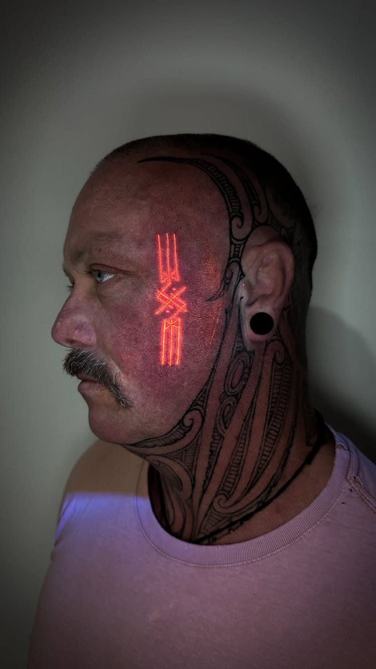 Man with black ink tattoos and a glowing red UV face tattoo.