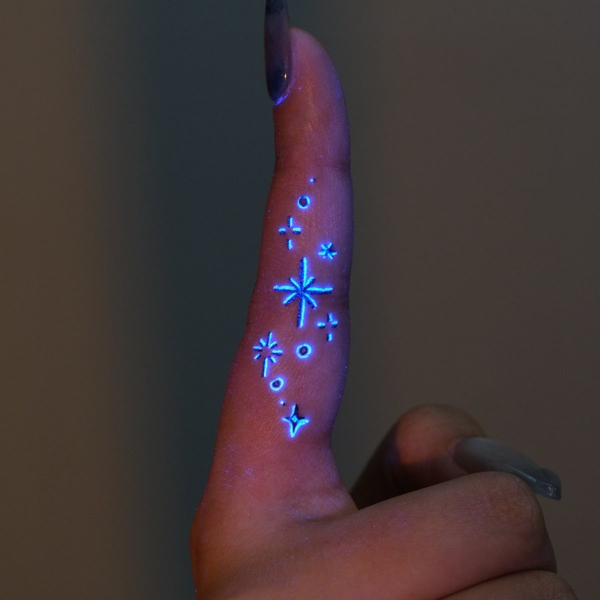 Glowing blue UV star tattoos running down a person's finger with long nails.