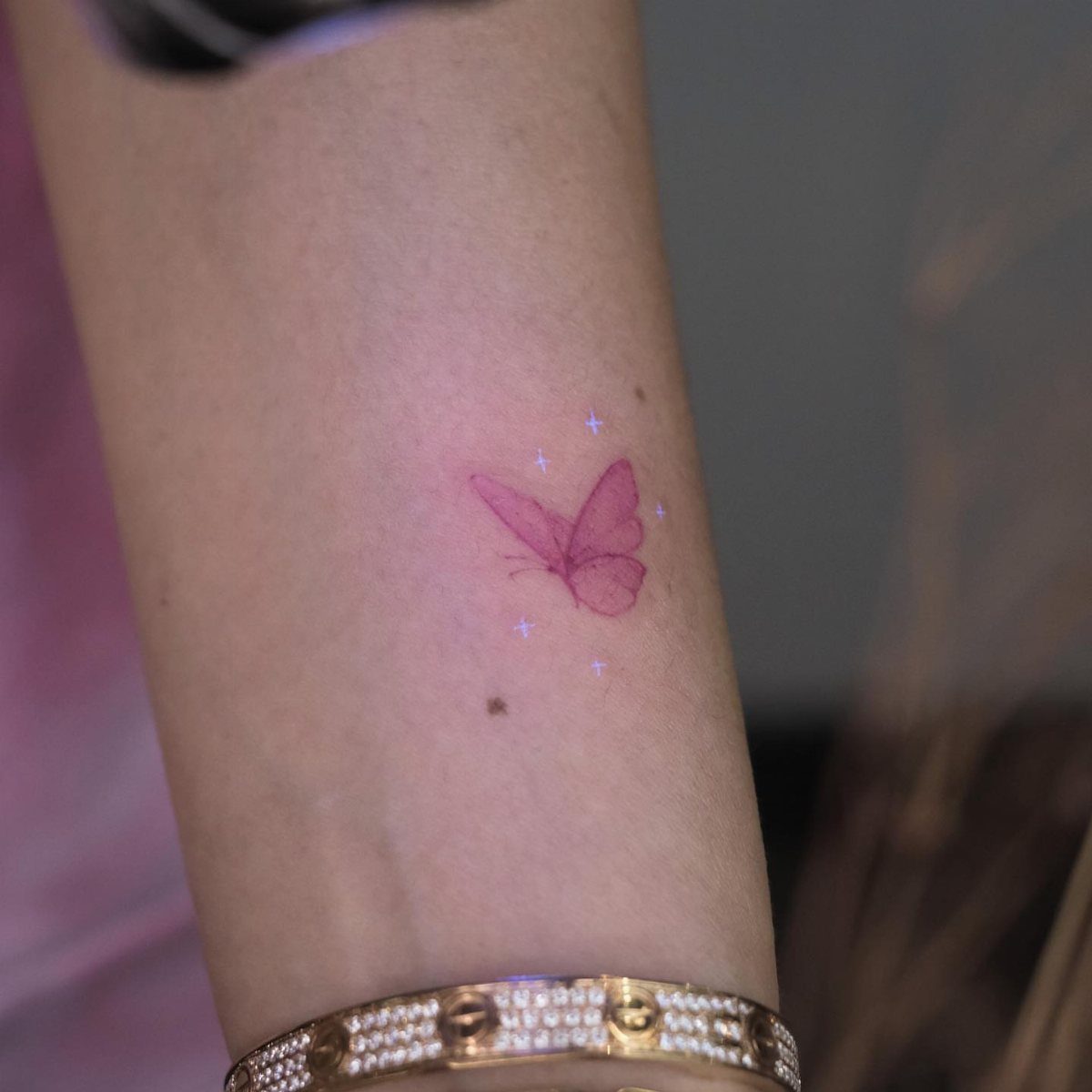 Glowing pink butterfly tattoo with stars on a person's forearm and gold bracelet.