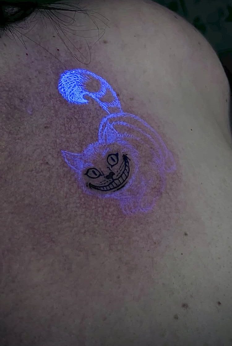 Cheshire Cat tattoo glowing purple and blue under UV light on a shoulder
