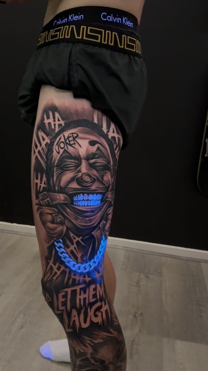 Joker clown tattoo on a thigh with glowing blue UV teeth and chain.